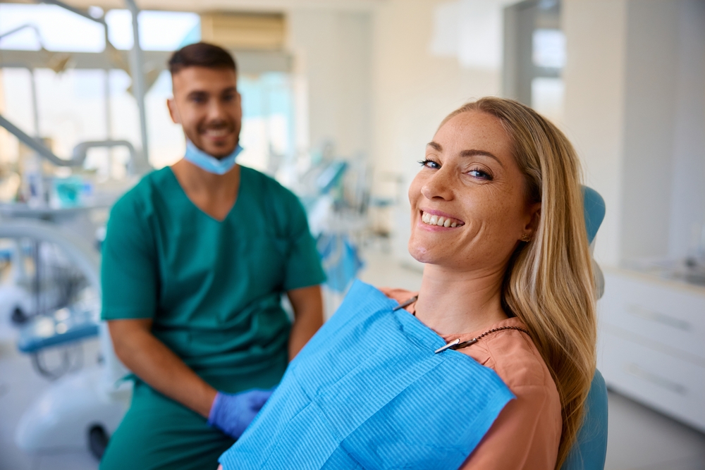 A woman in a dental chair smiles at the camera, wearing a blue bib. Behind her, a dentist in green scrubs and a mask sits and smiles. Dental equipment and bright lighting are visible in the modern clinic.