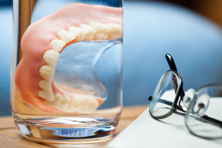 Full lower denture soaking in a glass of water on a bedside table, next to a pair of eyeglasses and a book—representing overnight denture care.