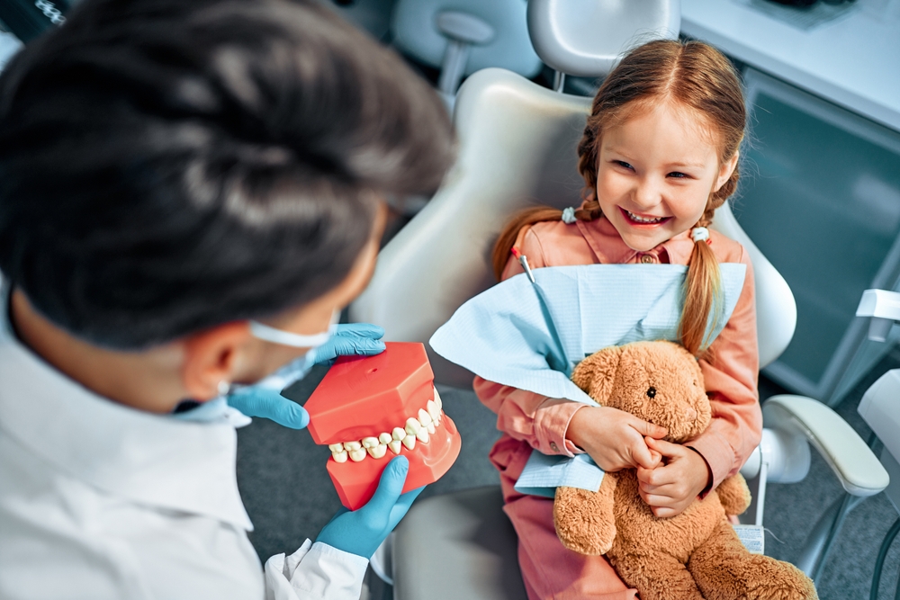 Photo of a senior man smiling genuinely at his dentist. The dentist is pointing to an x-ray that is displayed on a computer monitor behind them. No text on the image.